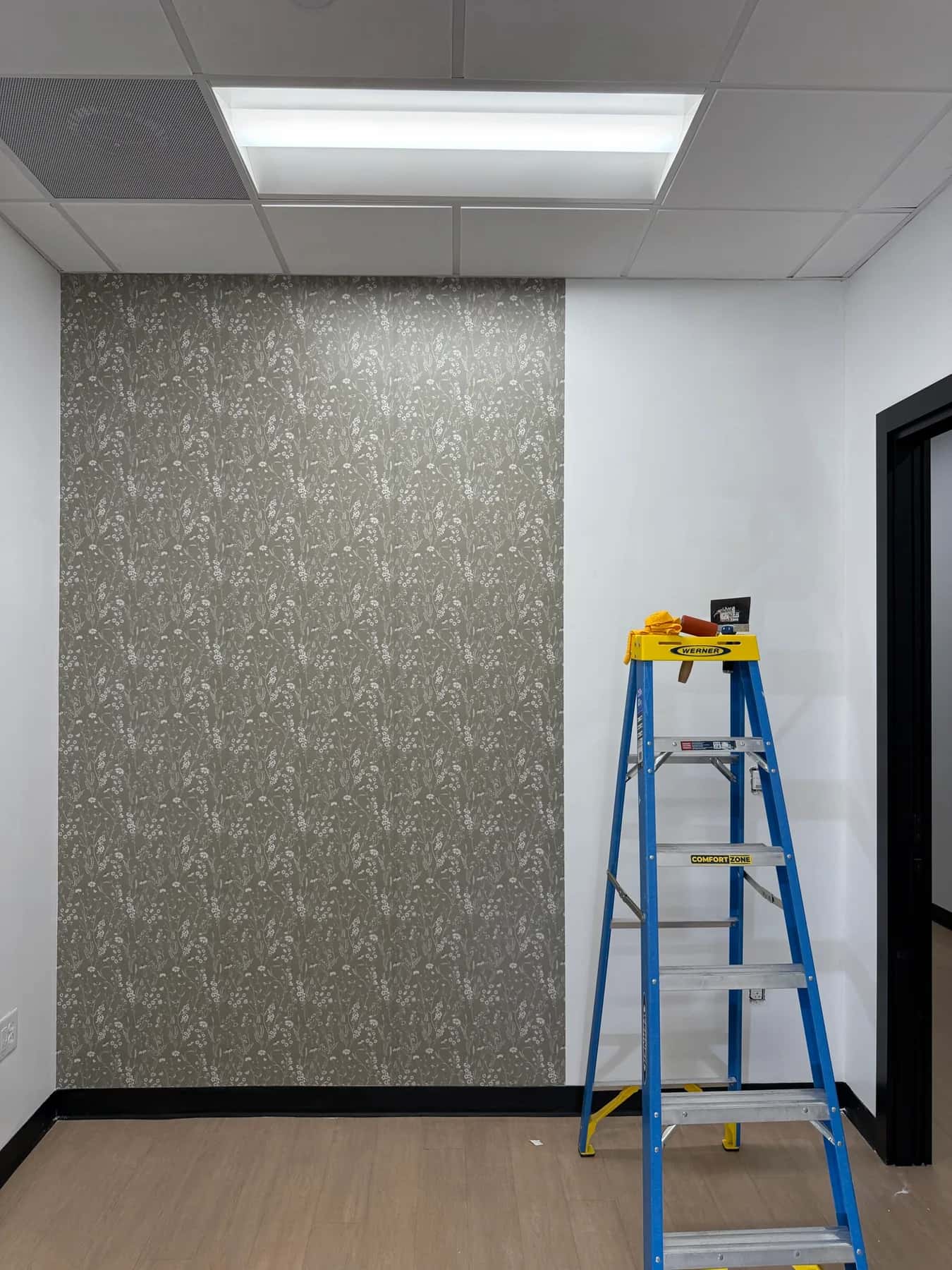 Sage green wildflower wallpaper covering half the accent wall, with a stepladder beside it