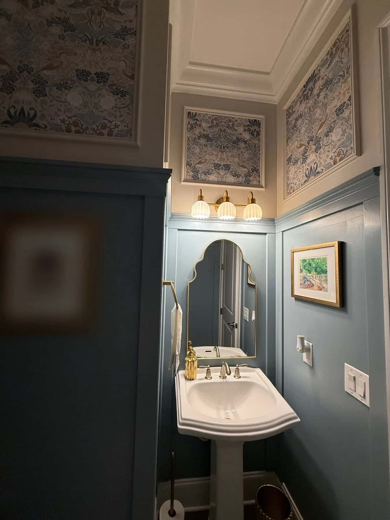 Wallpaper inside molding frames above a pedestal sink and arched mirror