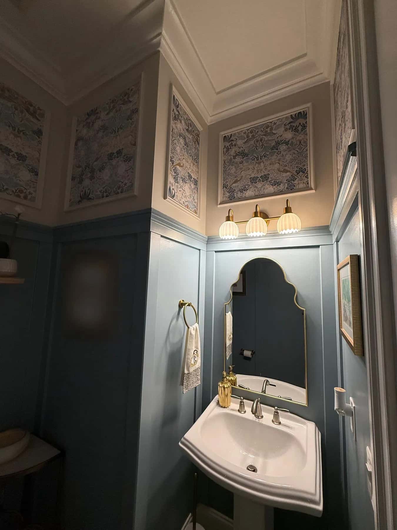 Framed wallpaper panels above a brass three-light sconce and arched mirror